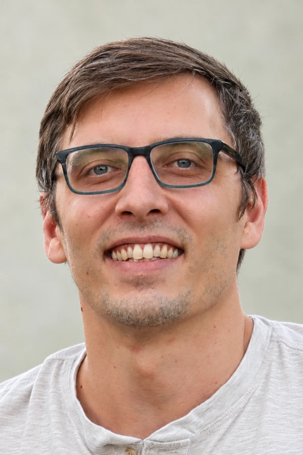 John Stanley smiling, wearing glasses and a light gray shirt against a neutral sage-gray background